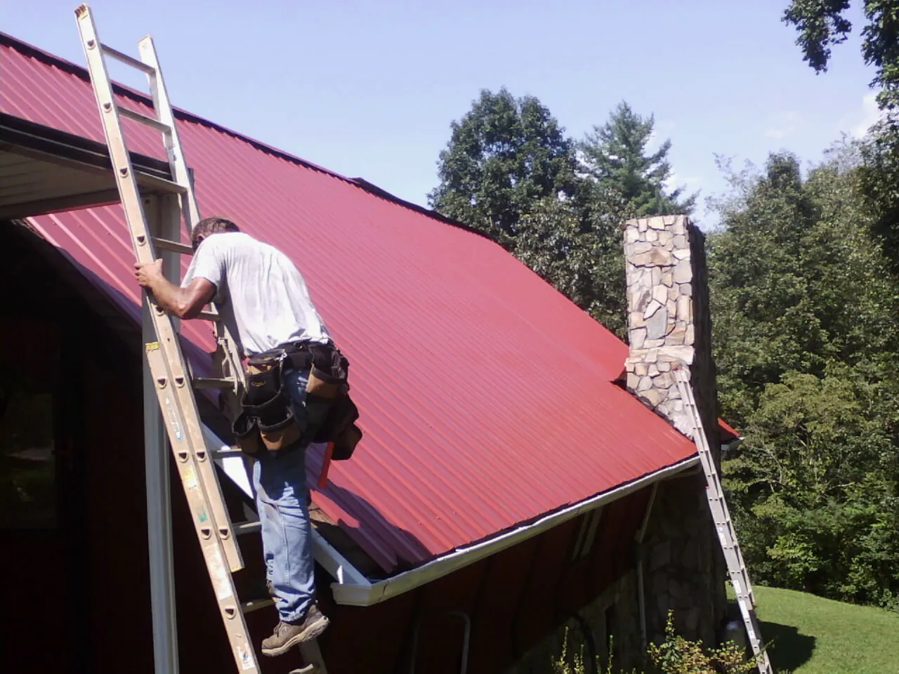 Roofer on a ladder beside a red metal roof and stone chimney — Soffit & Fascia Repair by Roofing Brothers in Beacon